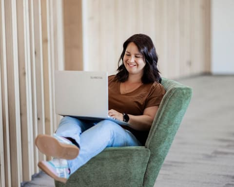 Elisabeth Zeilinger sitzt in einem grünen Ohrensessel, den Laptop auf dem Schoß, und tippt lächelnd.